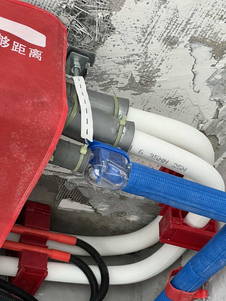 混凝土顶板下的空调接管近景，白色波纹软管经柔性接头接入灰色管件并用扎带锁紧，旁侧引出蓝色保温管，左上角露出红色标识布