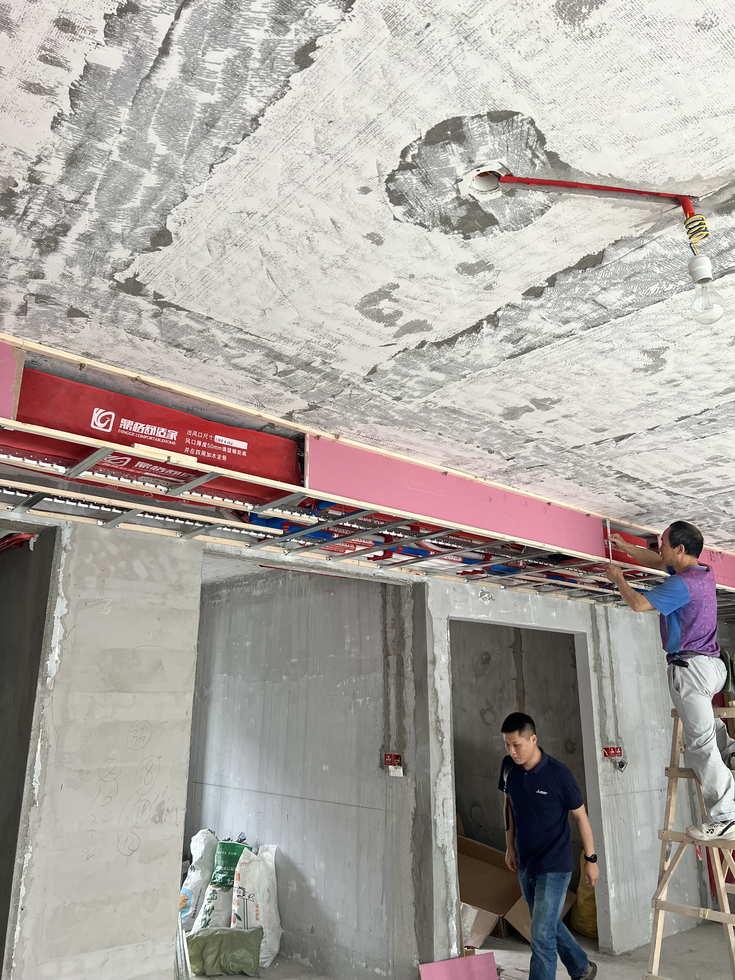 毛坯室内吊顶下方已就位的中央空调室内机，机身套有红色品牌保护罩，两名工人在梯子上进行吊顶与机器定位作业